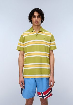 Giovane con capelli ricci che indossa una polo a righe verdi e gialle e pantaloncini blu con un disegno della bandiera rossa, bianca e nera.