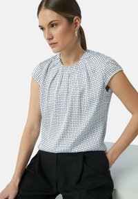 Kurzärmlige Bluse in Hellblau mit geometrischem Muster. Verfügt über einen gerafften Ausschnitt und einen weichen, strukturierten Stoff. Kombiniert mit schwarzen Hosen.