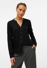 Blouse noire à manches longues avec un décolleté en V, dotée d'une fermeture à boutons et d'une poche poitrine simple. Texture lisse et léger éclat.