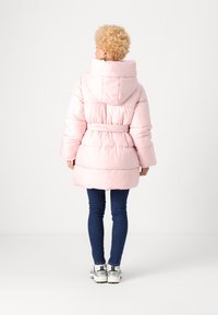 Cappotto puffo rosa con vita regolabile, grande cappuccio e design trapuntato. Indossato sopra jeans blu scuro e scarpe sportive argento. Vista posteriore.