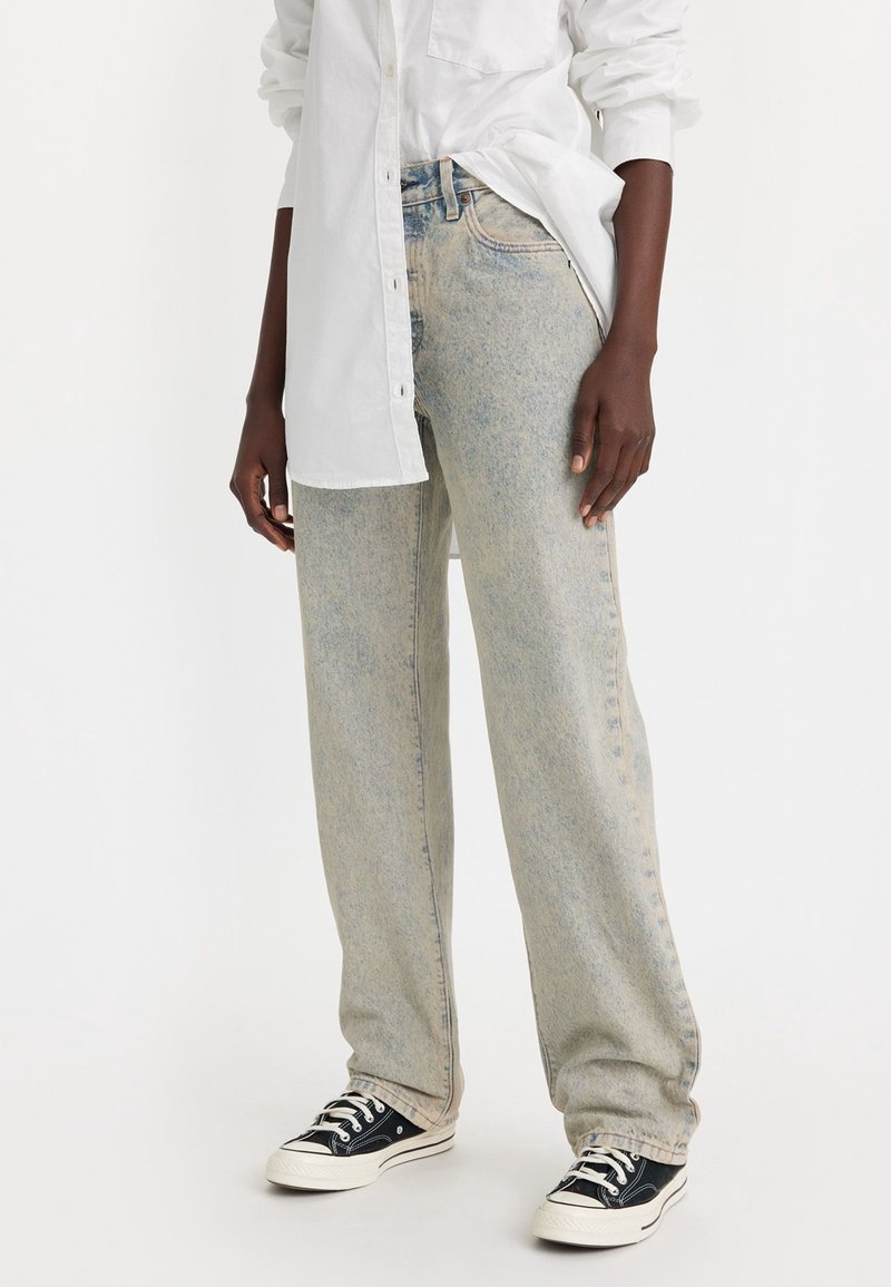 Jeans de mezclilla de color azul claro, ligeramente descoloridos, con un corte relajado, que presentan una cintura alta, cinco bolsillos y sutiles detalles de desgarre. Llevados con zapatillas negras.