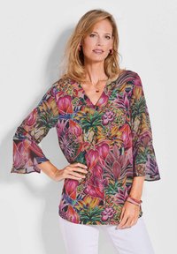 Transparente, blumige Bluse mit V-Ausschnitt und Glockenärmeln; bietet lebendige Muster in Pink, Lila und Grün; kombiniert mit weißen Hosen und Armbändern.
