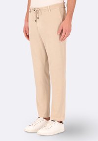 Pantaloni in misto cotone beige con vita regolabile tramite coulisse, gamba affusolata e tasche laterali, abbinati a sneaker bianche, presentati su uno sfondo neutro.