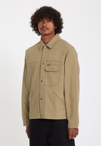 Homme aux cheveux bouclés portant une veste beige à boutons avec des poches poitrine et latérales, debout devant un fond blanc uni.