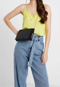 Mustan paljettikäsilaukku, jossa on teksturoitu, kuvioitu pinta. Mahdollisuus kantaa olkapäällä tai kädessä hopeaketjuhihnalla.