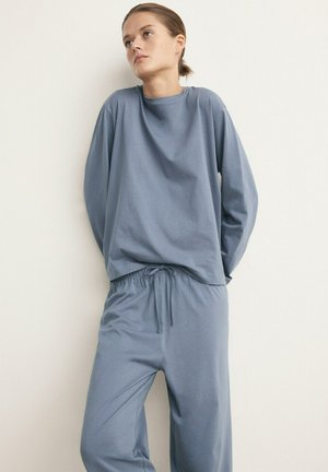 Junge Person lehnt an einer Wand und trägt ein locker sitzendes, blaues Langarmshirt und passende Hose mit Kordelzug, blickt leicht nach oben und zur Seite.