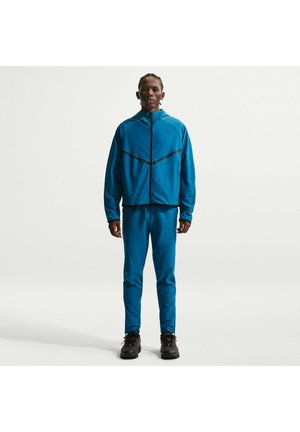 Uomo in piedi che indossa una giacca blu con zip e pantaloni abbinati con scarpe da trekking nere su uno sfondo bianco semplice.