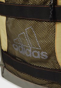 Sac à dos olive et gris avec un panneau en maille, un logo blanc, des bretelles noires et des accents de couture orange pour une touche de détail.