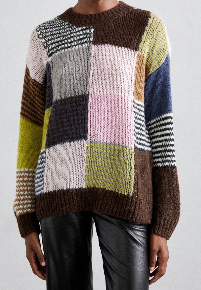 Pull en maille épaisse avec des motifs patchwork multicolores en marron, rose, jaune, bleu et noir, porté avec un pantalon en cuir noir.