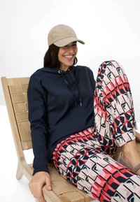 Felpa navy con coulisse, pantaloni rosa e rossi con motivo di forme geometriche, indossati con un berretto beige, seduto su una sedia di legno.