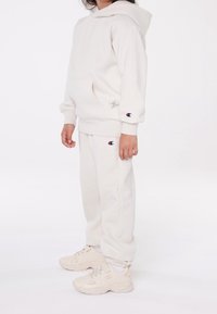 Barn som bär en off-white Champion hoodie, matchande sweatpants och beigefärgade sneakers, står mot en enkel vit bakgrund.