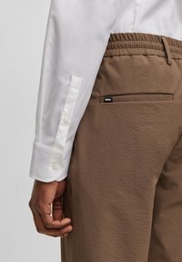 Bruna texturerade byxor med elastiskt midjeband, fickdetaljer och en svart "BOSS"-etikett. Vit skjorta med knäppta manschetter synlig på vänster arm.