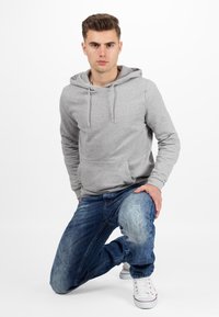 Sweatshirt à capuche grise avec une poche avant et des cordons de serrage, associé à un jean bleu délavé et des baskets blanches avec des accents rouges.