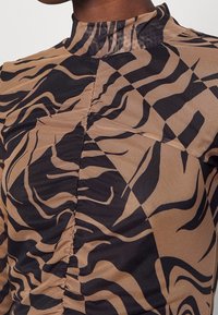 Haut à manches longues en tissu beige avec un motif de rayures de tigre noir. Présente un col montant et des détails froncés le long de la couture. Texture lisse.