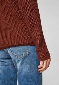 Textuurrijke, roestkleurige gebreide trui-mouw gecombineerd met lichtblauwe spijkerbroek, met subtiele vervaagde details en achterzakken.