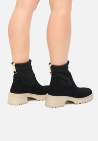 Czarne zamszowe botki za kostkę z prążkowanymi dzianinowymi mankietami, beżowymi obcasami na słupku oraz teksturowaną podeszwą, z pętlą jako ozdobą z tyłu.
