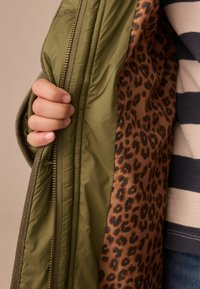 Grüne gesteppten Jacke mit einem braunen Leopardenmuster-Futter, ausgestattet mit einem goldenen Reißverschluss und strukturiertem Material. Eine Hand, die die Jacke greift.