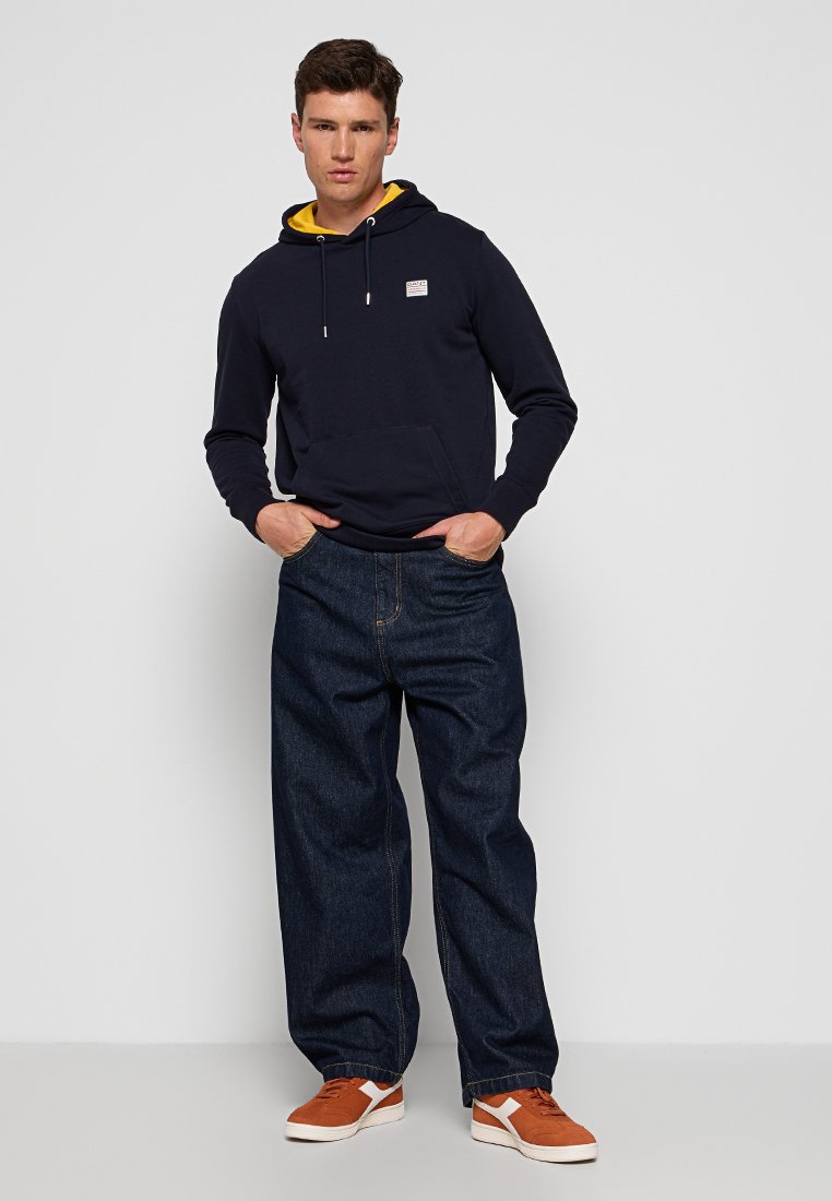 Hoodie bleu marine avec doublure jaune, poche frontale et patch logo, associé à un jean en denim sombre à jambes larges et des baskets marron avec des accents blancs.