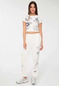 Giovane donna in t-shirt bianca corta e pantaloni jogger con stampe grafiche nere, in piedi con le mani rilassate, che indossa sneakers argentate su sfondo bianco.