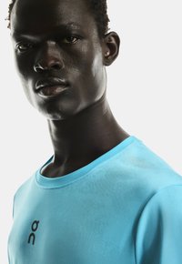 Bărbat tânăr cu pielea închisă la culoare, purtând un tricou sportiv de culoare albastră deschisă cu un logo negru "On", privind direct spre cameră pe un fond alb.