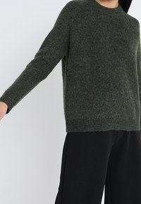 Dunkelgrüner, flauschiger Strickpullover mit langen Ärmeln, kombiniert mit weiten schwarzen Hosen vor einem schlichten hellen Hintergrund.