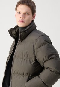 Jachetă puffer de culoare verde măslin închis, cu guler înalt, închidere cu fermoar și broderie discretă a logo-ului. Prezintă o textură netedă și lucioasă și bafe largi.