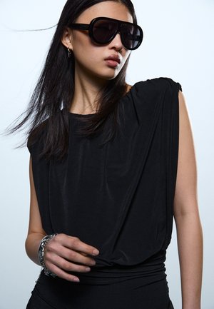 Mlajša ženska z dolgimi temnimi lasmi, ki nosi velike črne sončne očala, brezrokavnik črne barve in srebrno zapestnico, se pozira na enobarvnem ozadju.