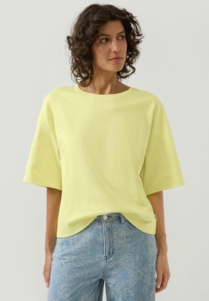 Kobieta o kręconych włosach, ubrana w luźną bladoróżową koszulę z krótkim rękawem oraz niebieskie dżinsy w kwiatowy wzór, patrząca na bok.