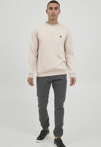 Lyse beige tekstureret sweatshirt med rund halsudskæring og et lille logo. Båret med mørkegrå tapered bukser og sorte sneakers.