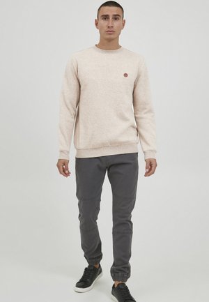 Világos bézs texturált pulóver kerek nyakrésszel és egy kis logóval. Sötétszürke tapered nadrággal és fekete sneakerrel párosítva.