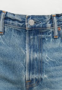 Vaqueros de denim azul con un lavado desgastado, ligera decoloración, herrajes de cobre y costuras naranjas a lo largo de la cintura y los bolsillos.
