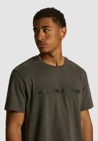 Szara bawełniana koszulka z okrągłym dekoltem i czarnym nadrukiem logo z przodu. Materiał ma gładką strukturę i krótkie rękawy.