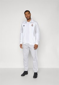 Giacca bianca con zip e accenti grigi, dotata di un logo e design a tre strisce. Abbinata a pantaloni grigi e scarpe da ginnastica nere.