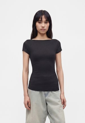 Jeune femme aux longs cheveux noirs portant un haut noir moulant à manches courtes et un pantalon ample gris clair sur un fond clair uni.