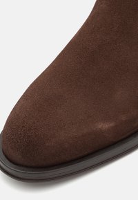 Bout de chaussure en daim marron avec forme arrondie et semelle en caoutchouc noire sur fond clair.