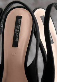 Mustat mokkanahasta valmistetut slingback-korkokengät, joissa on sileä vaaleanpunainen nahkasisäpohja. Kengissä on mustat tarrat, joissa lukee valkoisella kirjasimella "TABITHA SIMMONS" ja "NEXT".