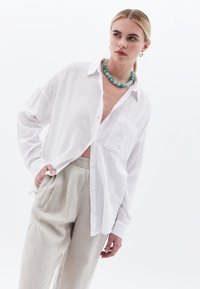Camisa blanca de botones en tejido texturizado con un bolsillo en el pecho, combinada con pantalones beige claros. Accesorios con un collar de cuentas turquesas.