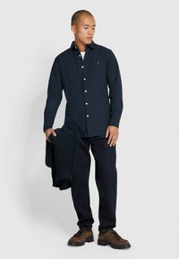 Granatowa koszula zapinana na guziki z długim rękawem i białymi guzikami, w połączeniu z ciemnymi dżinsami. Trzyma ciemny sweatshirt, ma na sobie brązowe buty.