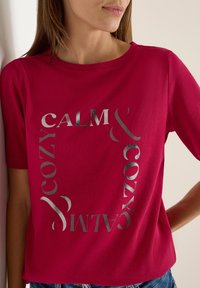 Fuksjowa koszulka z krótkim rękawem z srebrnym, odbijającym światło napisem "CALM & COZY" w okrągłym układzie. Wykonana z miękkiej bawełny.