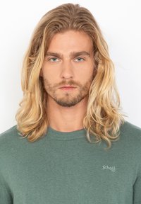 Zielony sweter dzianinowy z okrągłym dekoltem. Posiada subtelne logo na klatce piersiowej i miękką teksturę. W tle długie, falujące blond włosy.