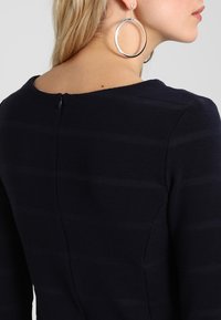 Vestido azul marino con rayas horizontales texturizadas, con cuello redondo y cierre de cremallera en la parte posterior. Aros de plata grandes visibles.