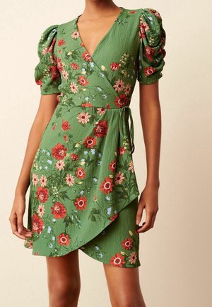 Frau trägt ein grünes Wickelkleid mit roten und pinkfarbenen Blumenmustern und puffigen kurzen Ärmeln.