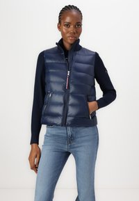 Gilet imbottito navy con trapuntatura, colletto alto, chiusura con zip e tasche laterali. Indossato sopra un maglione scuro e jeans azzurri chiari.