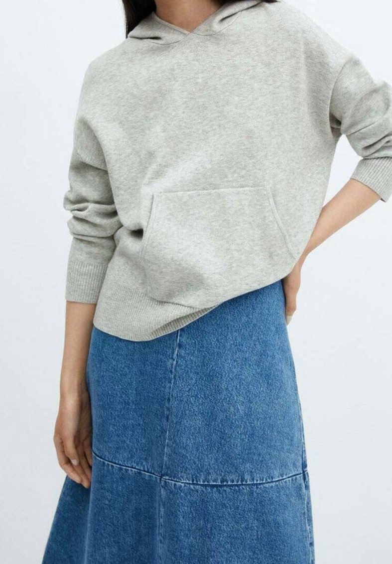Femme portant un sweat à capuche gris clair avec poche avant et une jupe en jean bleu en forme trapèze, debout avec une main sur la hanche.