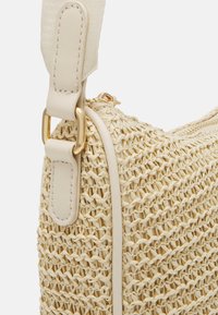 Pleciona torebka ze słomy w kolorze kremowym, wyposażona w gładkie skórzane wykończenia, regulowany pasek i złote elementy ozdobne. Teksturowana powierzchnia z misternymi wzorami.