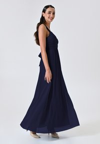 Abito maxi blu navy con scollatura arricciata, spalline sottili, tessuto fluido e un fiocco sul retro. Indossato con tacchi nudi.