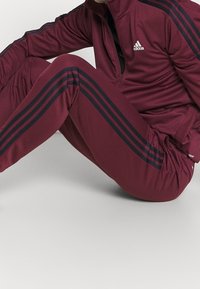 Personne portant un survêtement Adidas bordeaux avec des bandes noires, assise avec les jambes pliées sur un fond clair uni.