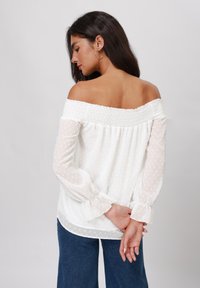 Off-the-shoulder white blouse with textured dot pattern, smocked neckline, and sheer long sleeves featuring gathered cuffs. Worn with blue jeans.