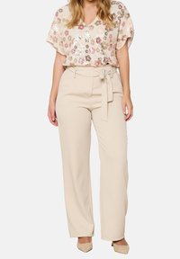 Donna che indossa pantaloni beige a vita alta con cintura da annodare, una blusa a sequenze floreali e tacchi a punta beige, in piedi contro uno sfondo bianco.