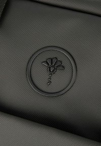 Sort struktureret materiale med en cirkulær præget logo, der har et blomsterdesign i midten, uden yderligere accenter eller synlige funktioner.